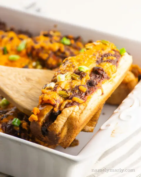 A vegan baked hot dog is held on a spatula over the rest of the casserole dish.