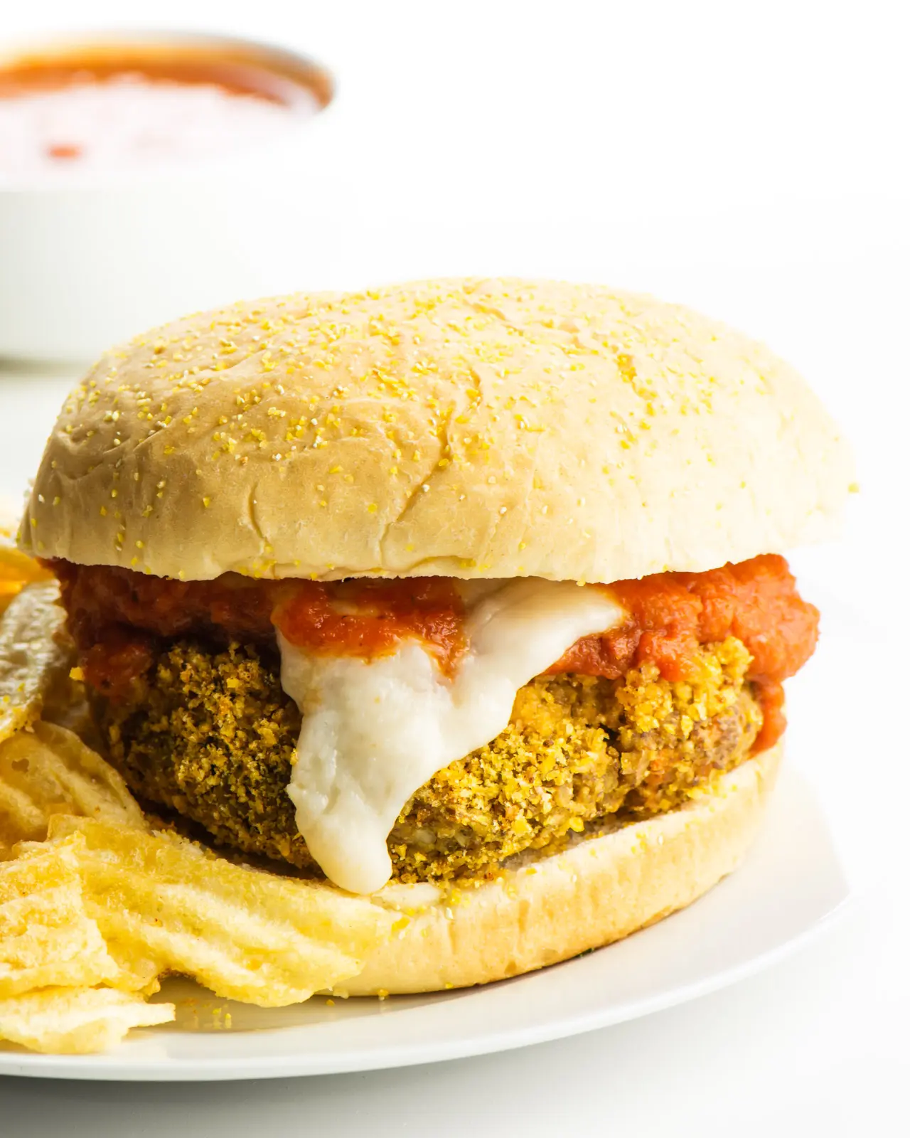 A vegan parmesan sandwich sits on a white plate with chips beside it and marinara sauce in a bowl behind it.