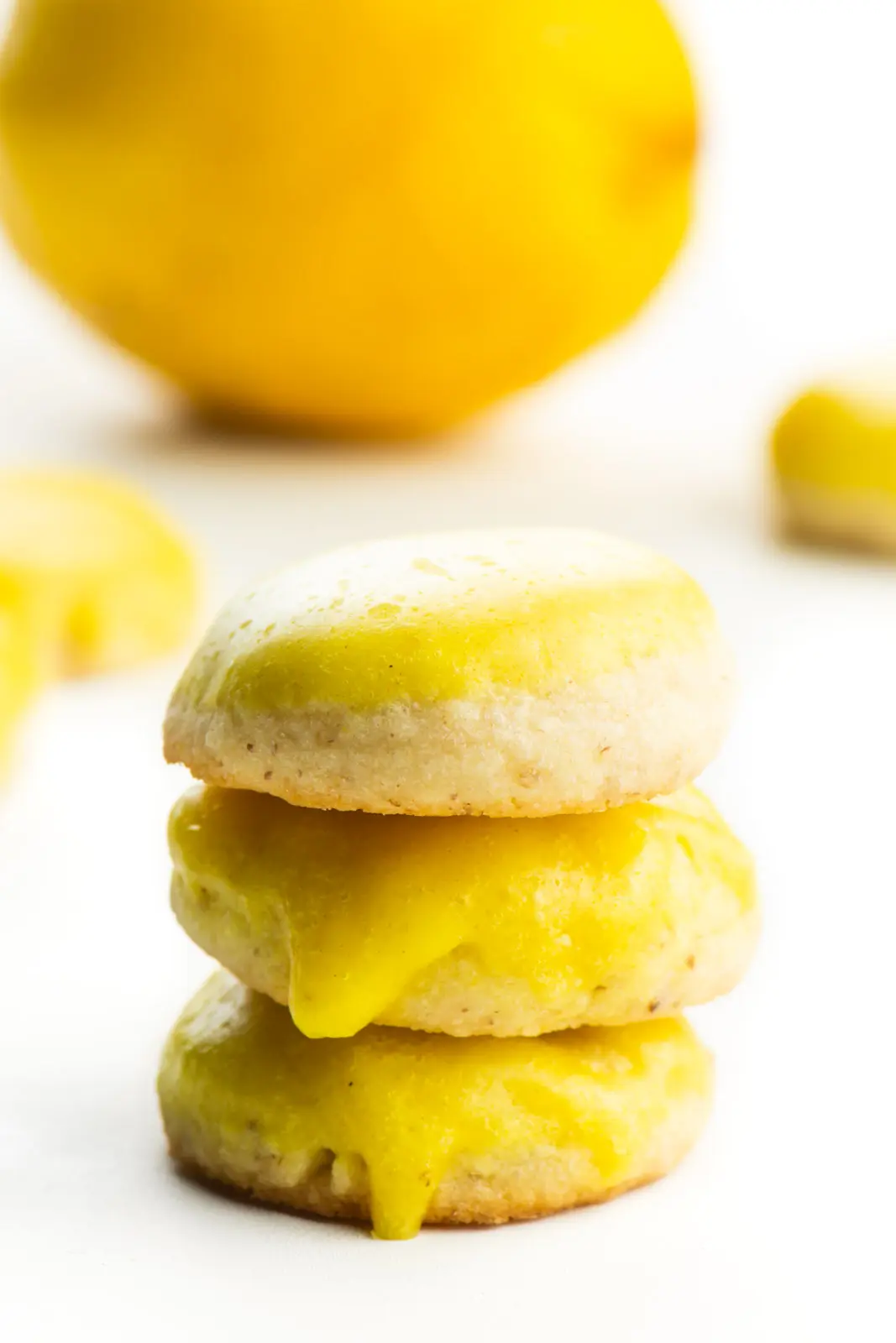 The vegan lemon cookies are stacked on top of each other with more cookies behind it and a lemon.