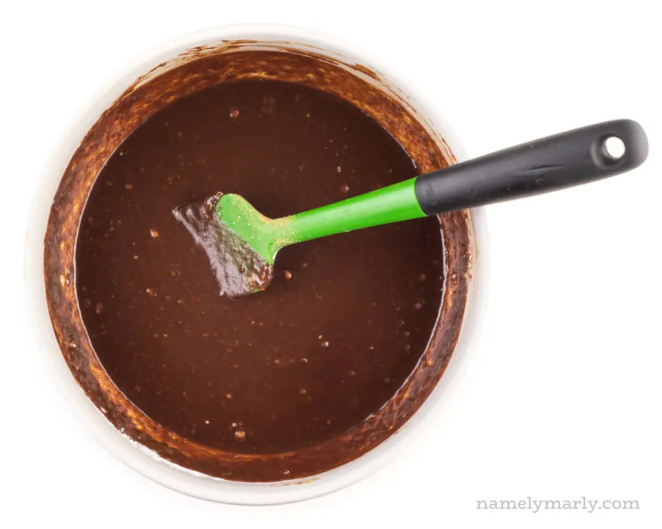 A chocolate mixture is in a glass bowl with a green spatula.