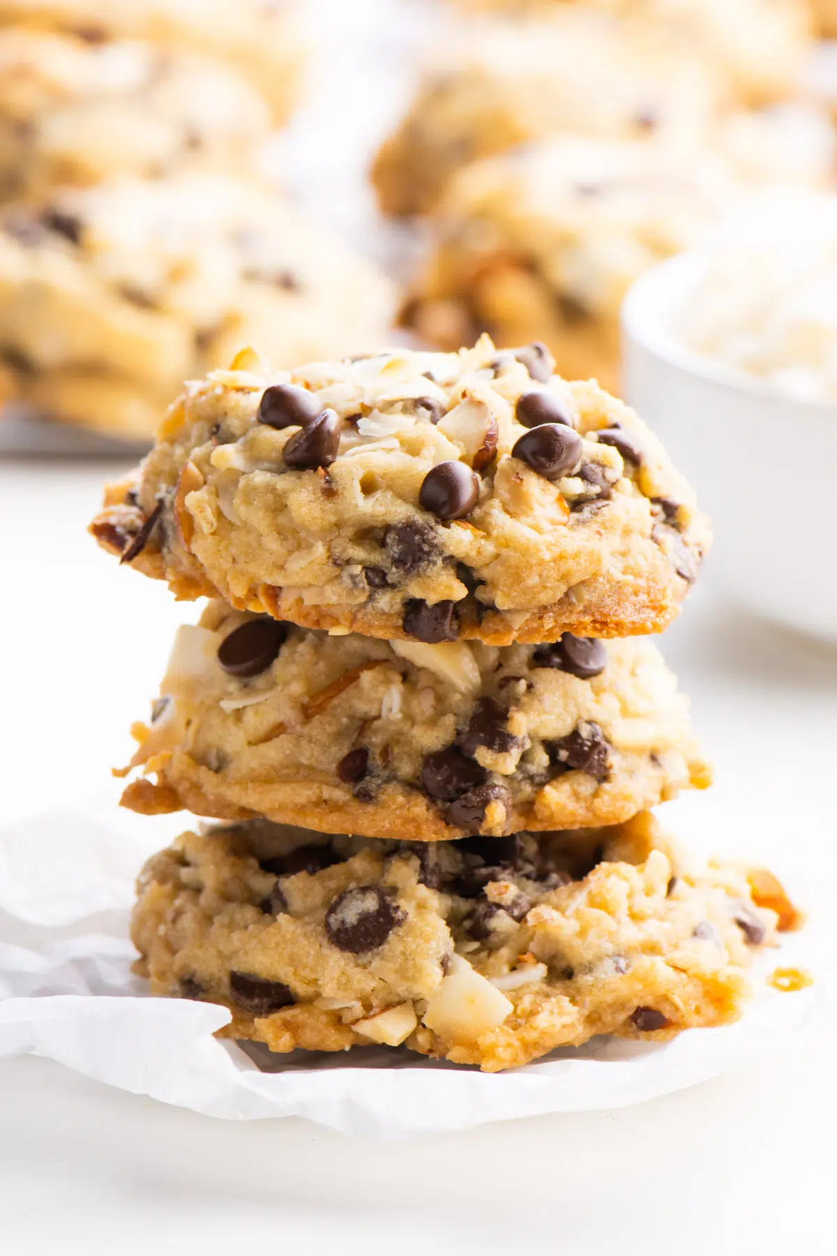 A stack of three almond joy cookies sits in front of a baking rack with more vegan cookies.