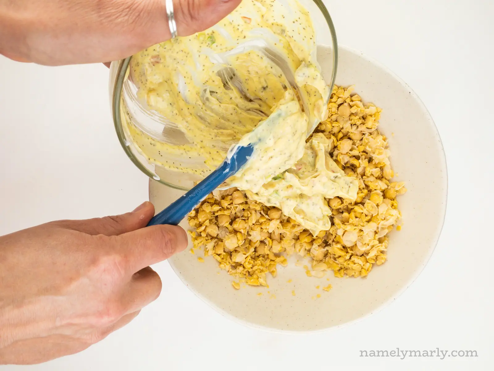 A hand holds a glass bowl over mashed chickpeas and uses a spatula in the other hand to spread it over the chickpeas.