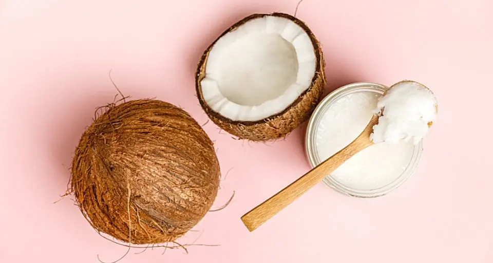 A coconut is cut in half, with one half showing the brown outer crust and the other half showing coconut inside. There is a spoon sitting on a jar of coconut oil next to the coconuts.