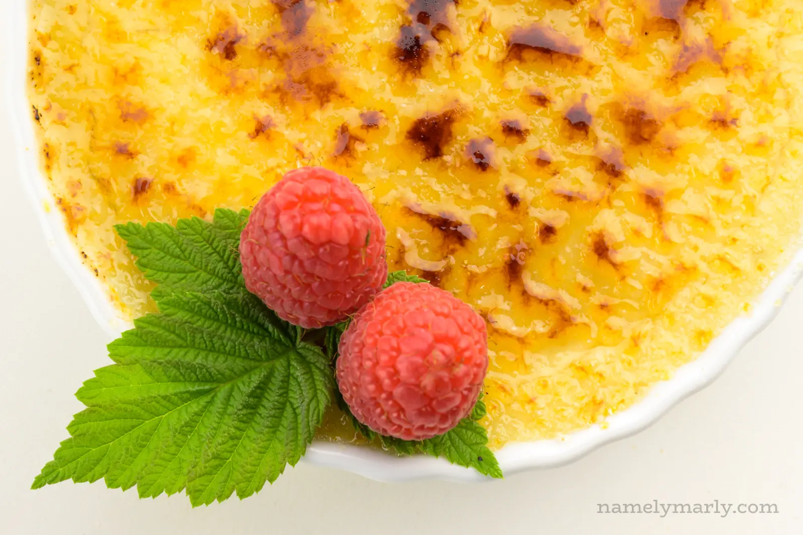 A close-up shot of creme brûlée in a ramekin with burnt sugar on top and raspberries on the side.