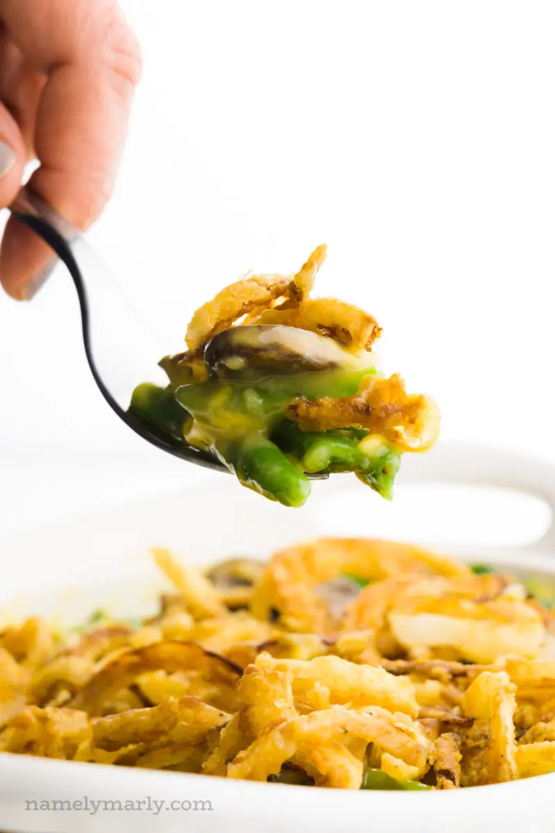 A hand holds a fork full of vegan green bean casserole with the rest of the dish behind it.