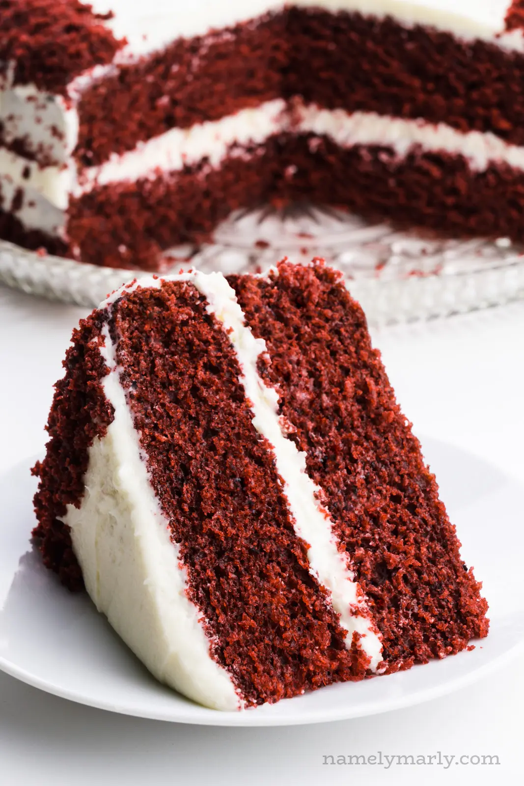 A slice of red velvet cake on a plate in front of the rest of the cake.