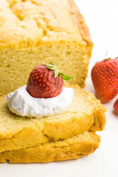 A loaf cake has two slices cut out of it and a dollop of whipped cream is on top with strawberries on top and beside it.