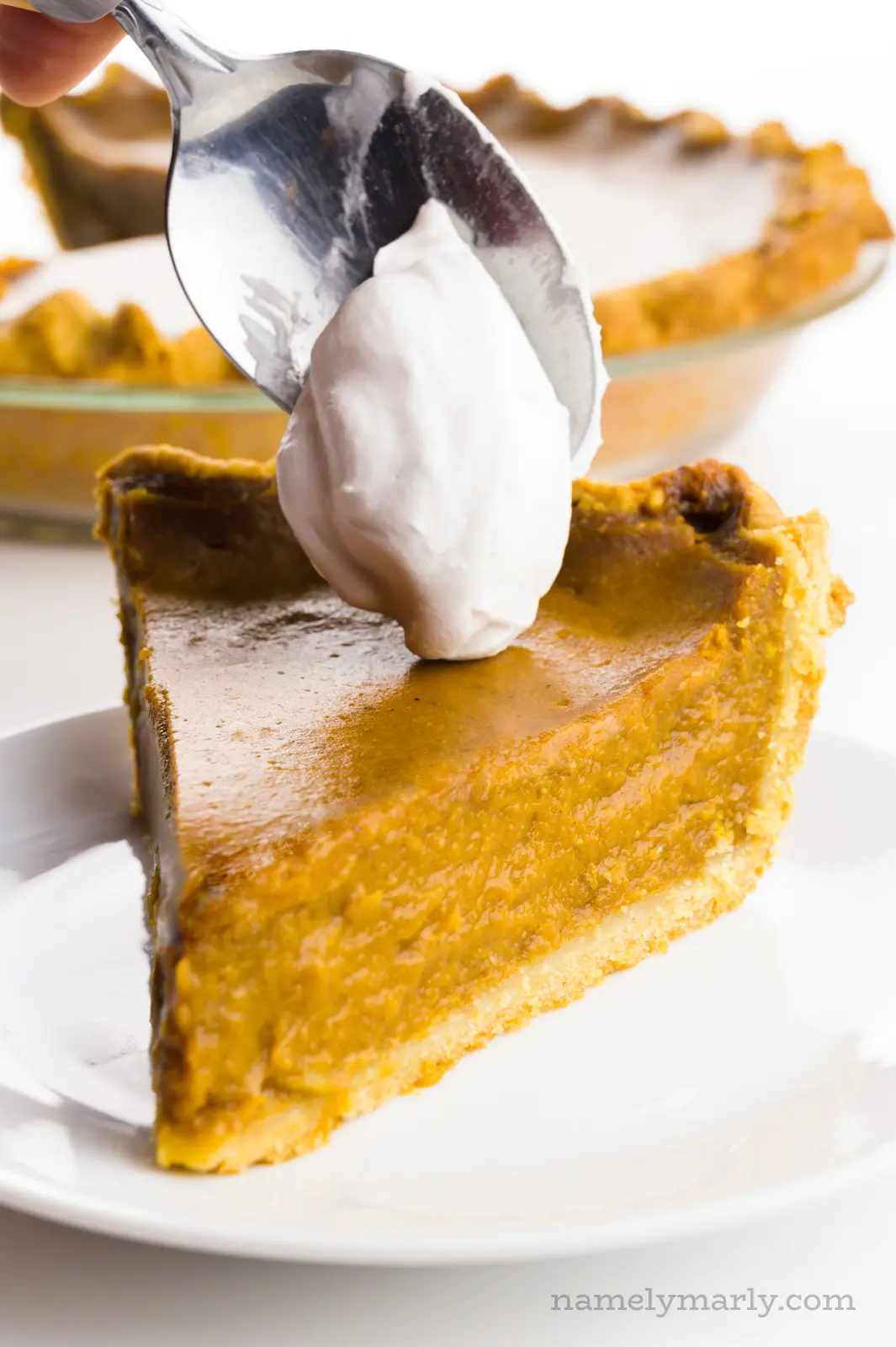 A hand holds a spoon dropping whipped cream on a slice of pumpkin pie.