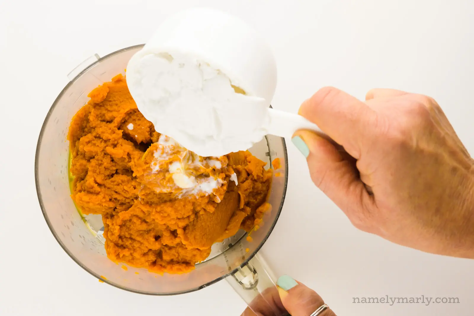 Coconut milk is being poured into a food processor bowl full of pumpkin puree.