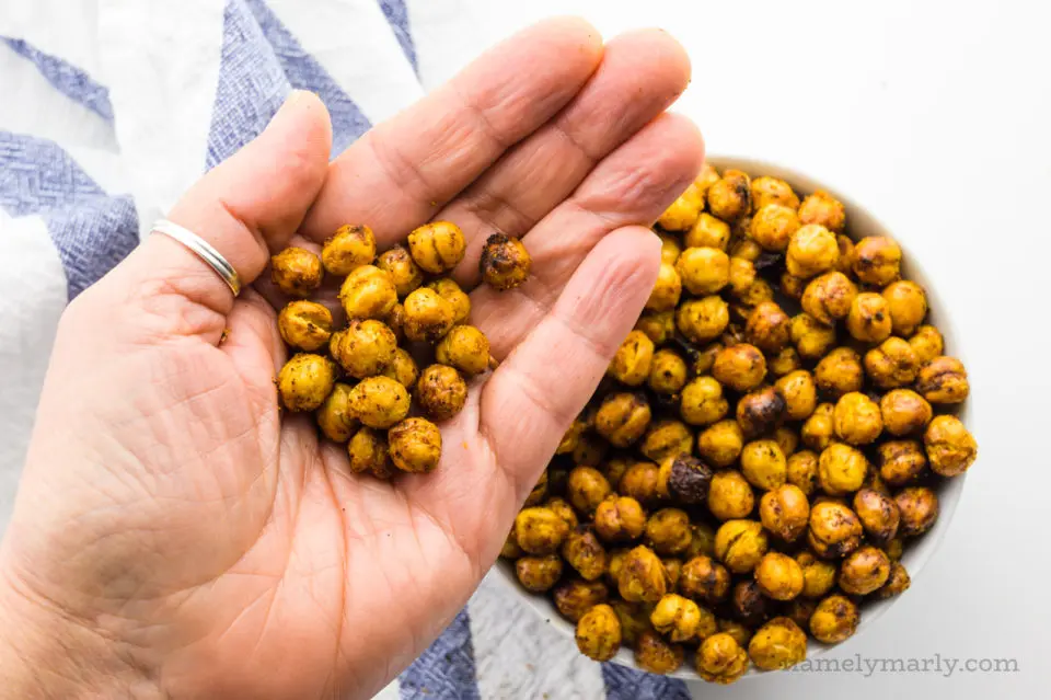 A hand holds several air fryer chickpeas, with a bowl of them behind it.