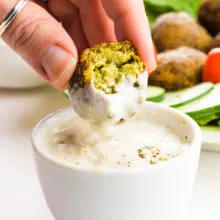A hand holds an falafel and is dipping it in a white sauce.