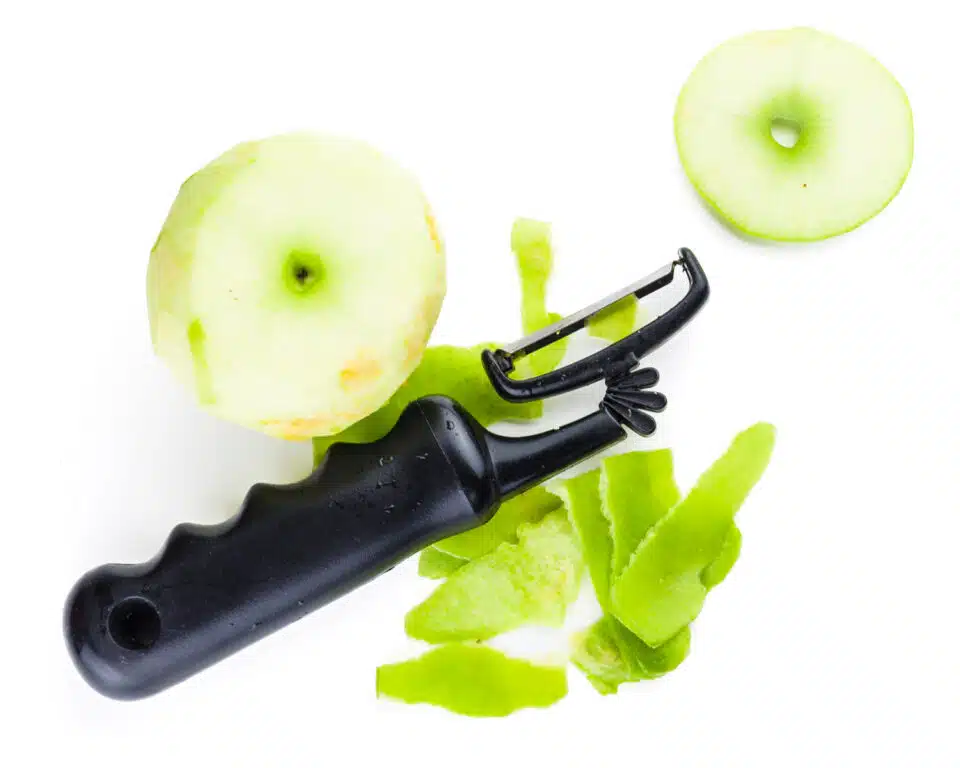 A vegetable peeler sits next to an apple with the top chopped off. Several peels from the apple sit beside it.