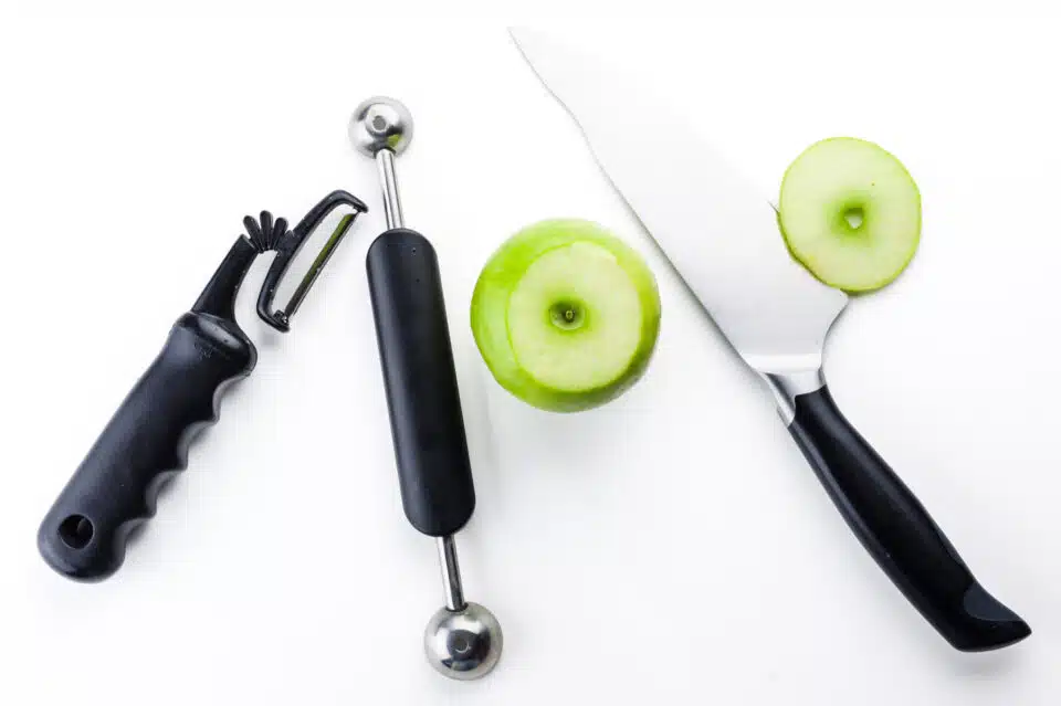 A green apple has the top cut off, and it's sitting next to a butcher's knife, a vegetable peeler, and a melon baller.