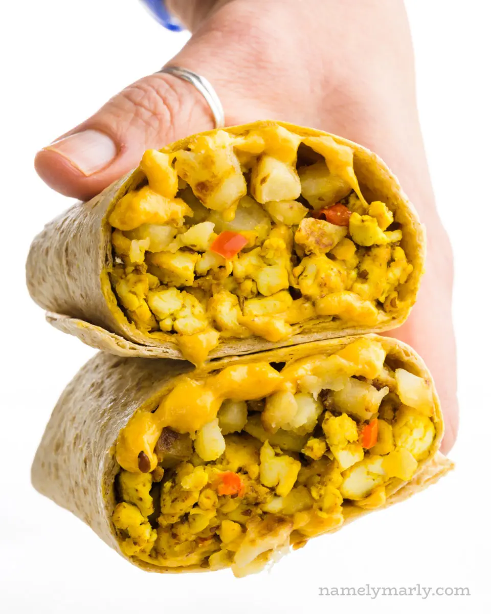 A hand holds two halves of a breakfast burrito, showcasing the ingredients.