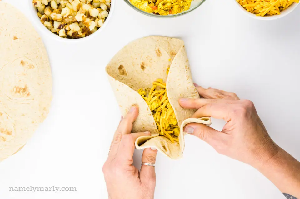 Two hands grasp a tortilla, rolling it into a breakfast burrito. There are other ingredients around the tortilla.