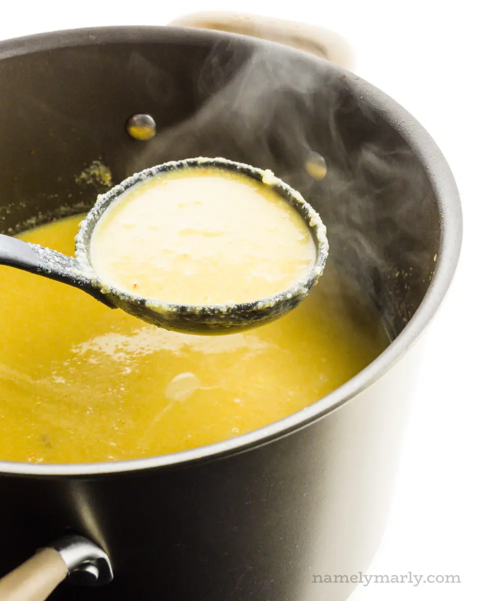A ladle is full of soup, hovering over the rest of the pot of soup. Steam rises above the hot soup.