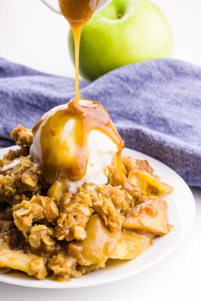 A plate holds vegan apple crisp with ice cream on sauce. Caramel sauce is being poured over the top. A blue napkin and green apple is behind it.