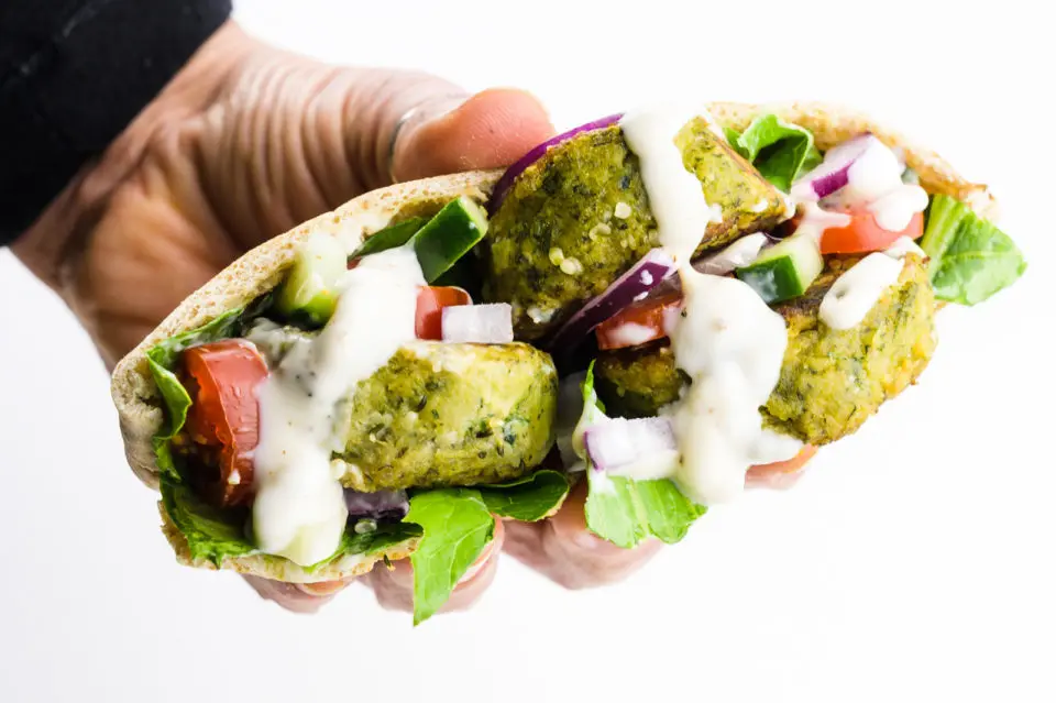 A hand holds a baked falafel sandwich with greens and sauce.