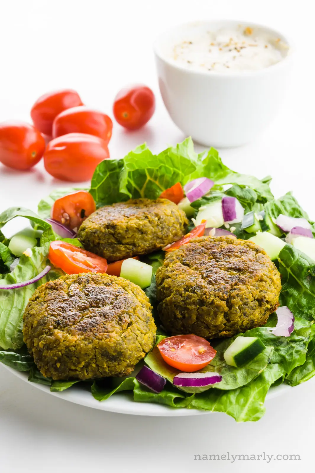 A salad is topped with baked falafel and tomatoes. There are more tomatoes behind it and a bowl full of tahini sauce.
