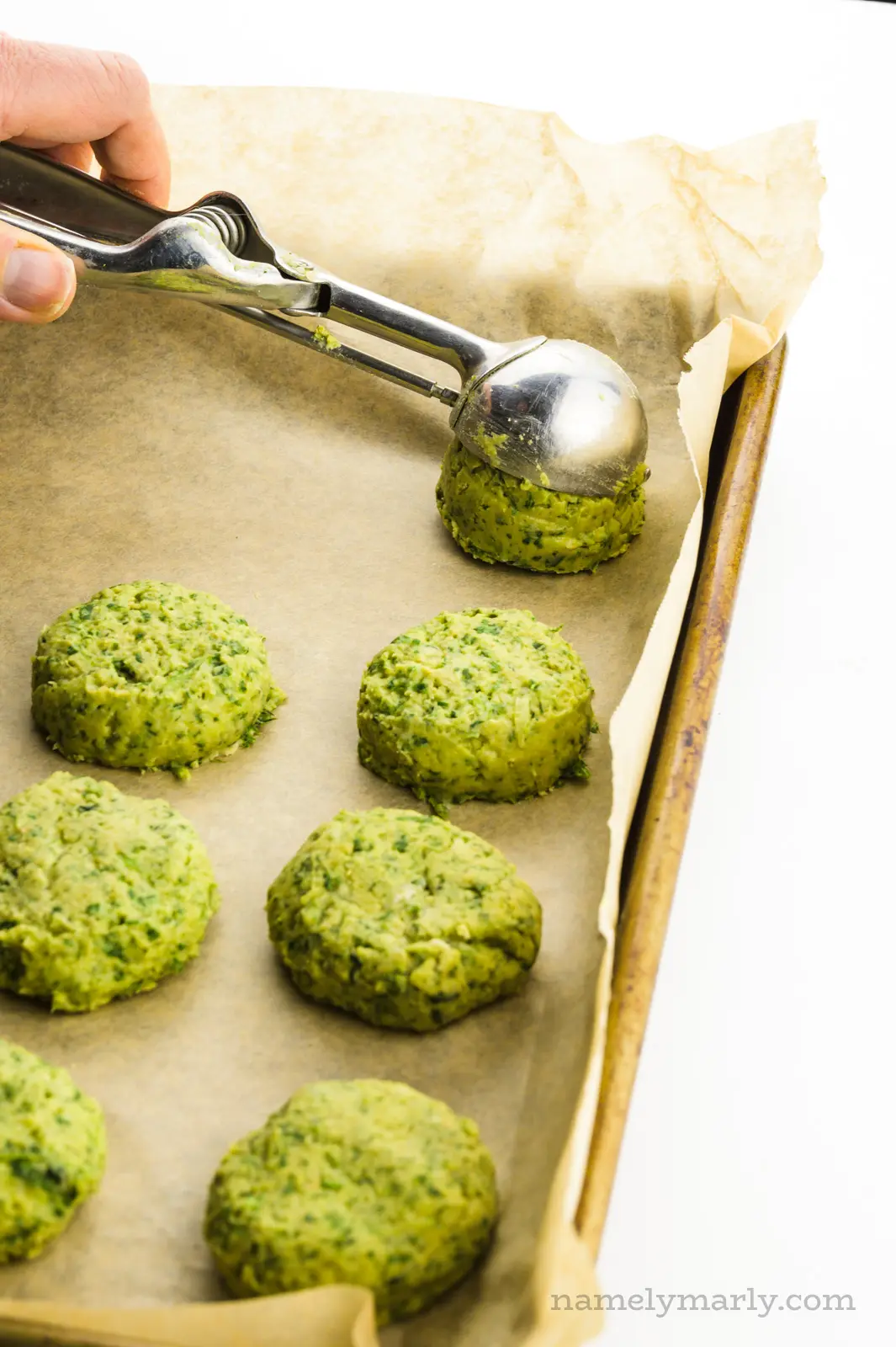 A cookie scoop is measuring falafel batter onto a baking sheet lined with parchment paper.