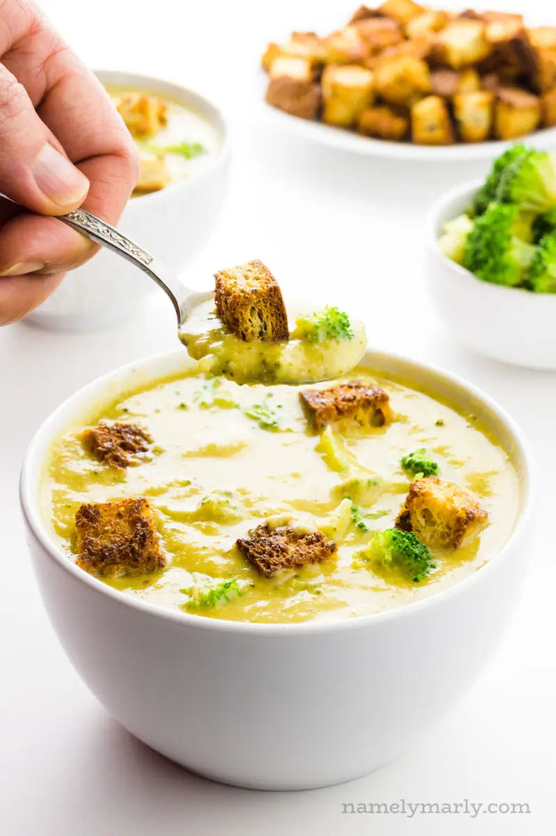 A hand holds a spoonful of soup over the bowl. There are bowls of steamed broccoli and croutons behind it.