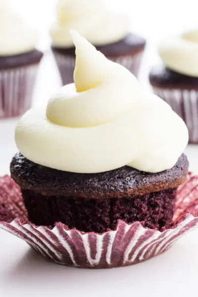 A red velvet cupcake is topped with vegan cream cheese frosting. There are more cupcakes behind it.
