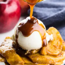 Caramel sauce is being drizzled over ice cream atop baked apple slices. There is an apple and a blue kitchen towel behind it.