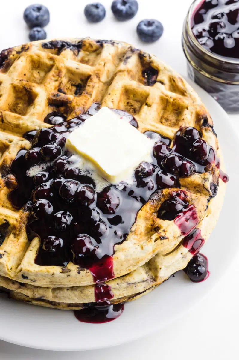 A stack of blueberry waffles are topped with blueberry sauce and melty butter. A jar with more blueberry sauce is behind it.