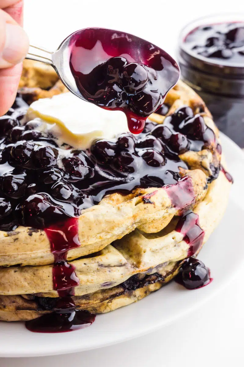 A spoon full of blueberry sauce hovers over blueberry waffles covered in sauce and vegan butter.