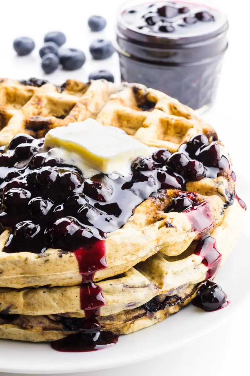 A stack of blueberry waffles has vegan butter on top and some blueberry sauce. There are fresh blueberries and a bowl of more blueberry sauce behind it.