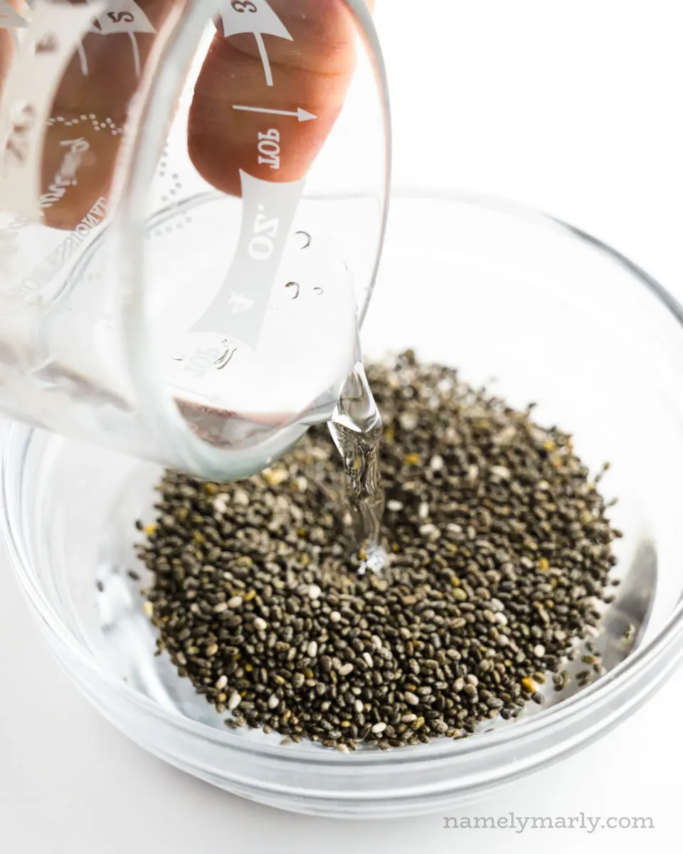 A hand holds a glass measuring cup with water and is pouring it into a bowl with chia seeds.