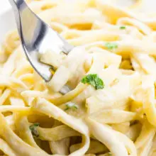 A fork dips into a bowl of fettuccine covered with vegan alfredo sauce and parsley sprigs.