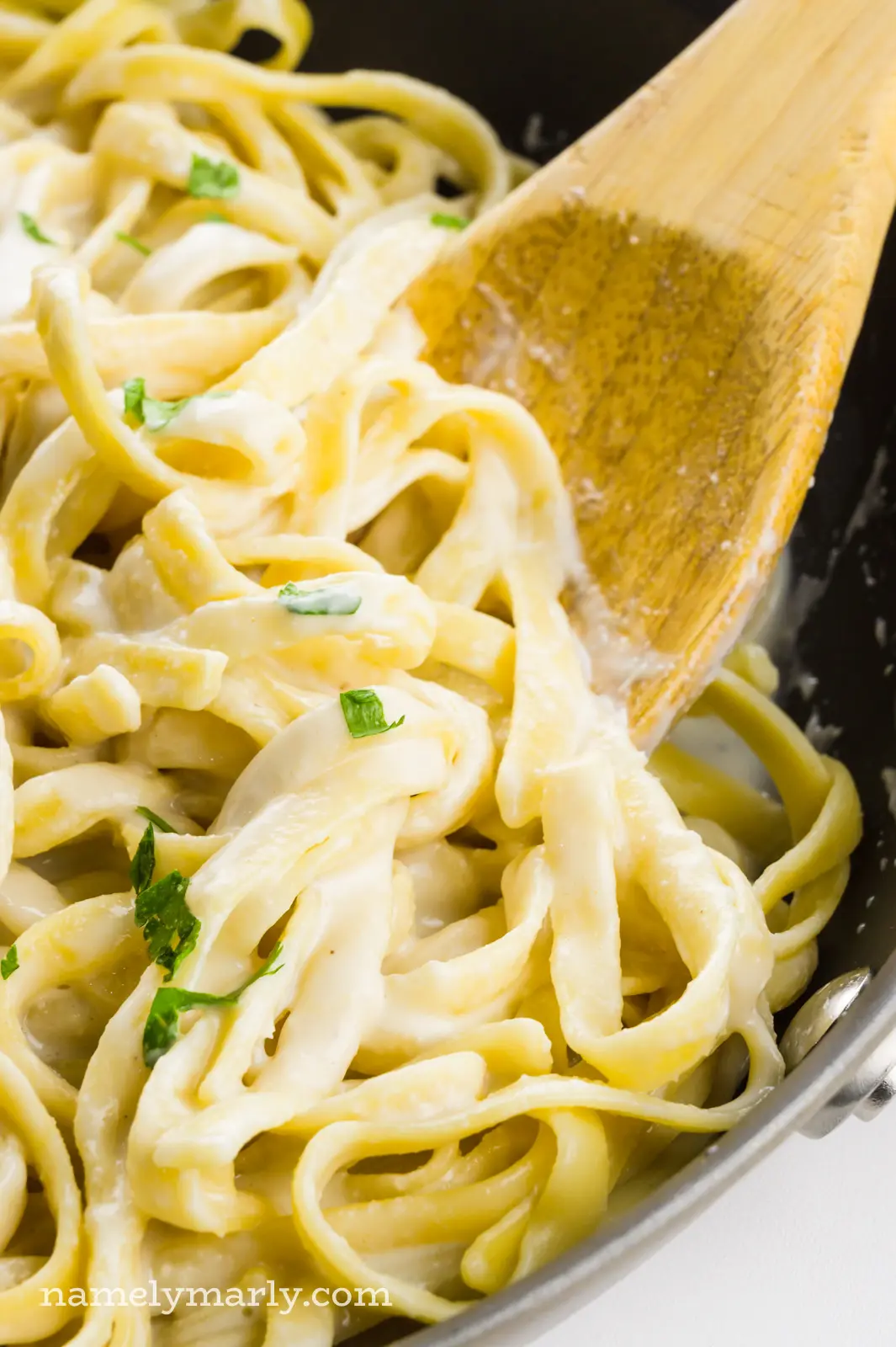 A wooden spoon scoops out fettuccine alfredo from a skillet.