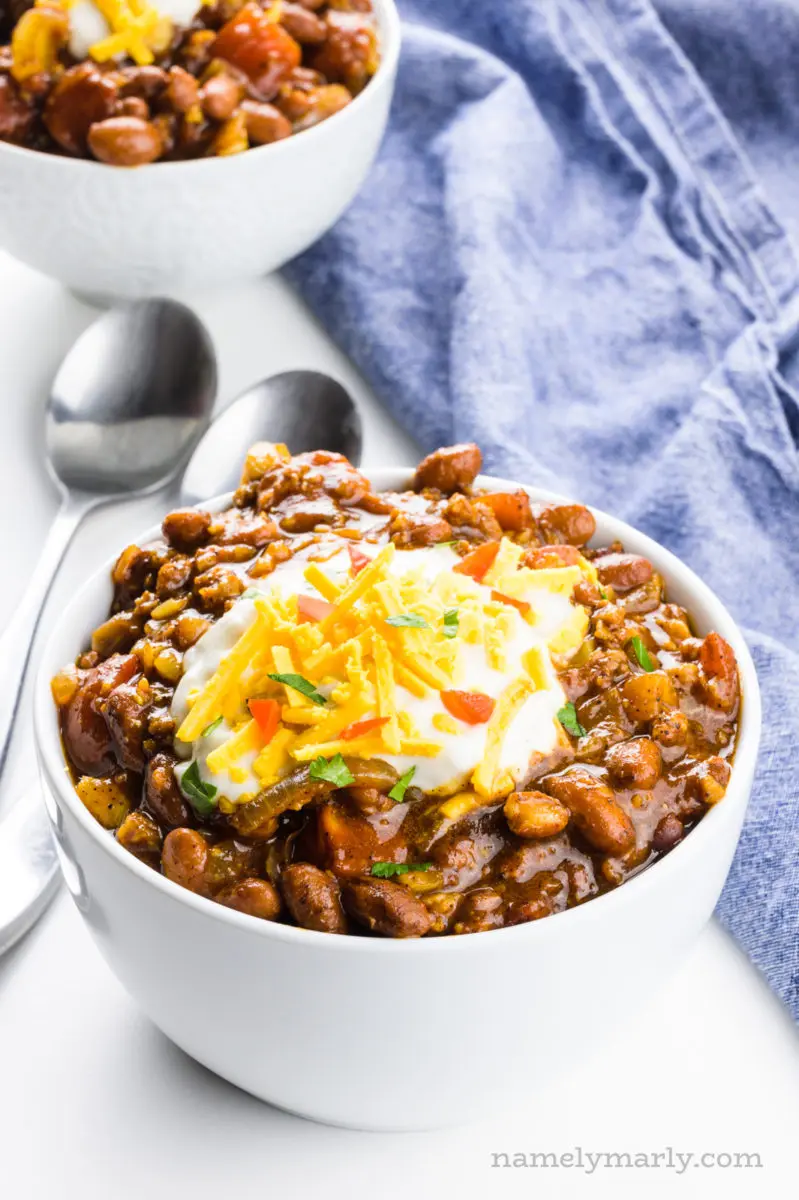 Two spoons sit between two bowls of chili, both topped with vegan sour cream and other toppings. A blue kitchen towel is beside it.