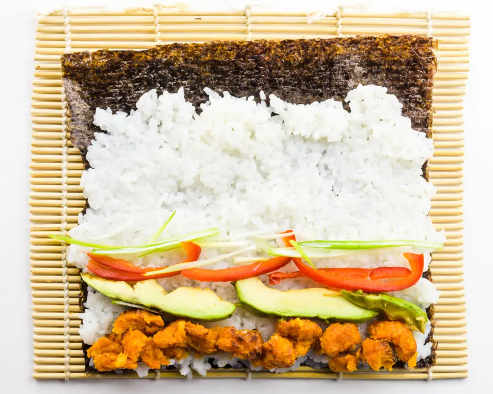 A nori sheet is on a bamboo mat with ingredients on top like rice, sweet potato pieces, avocados, and more.