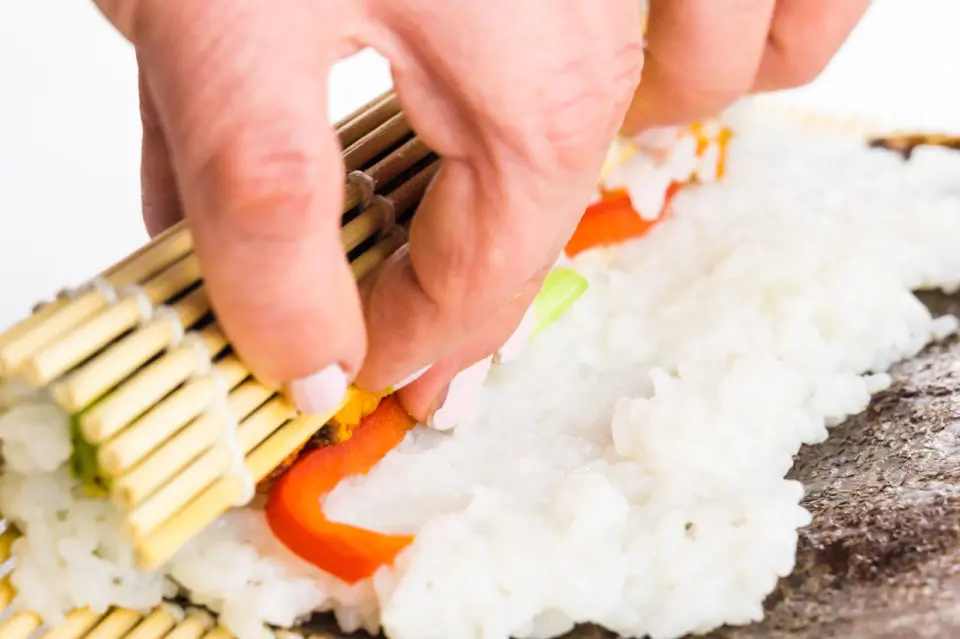 Hands are using a bamboo mat to roll up a sheet of nori with sushi ingredients on top.