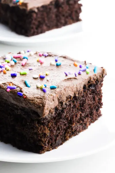 A slice of chocolate cake has frosting and sprinkles on top. Another slice sits behind it.