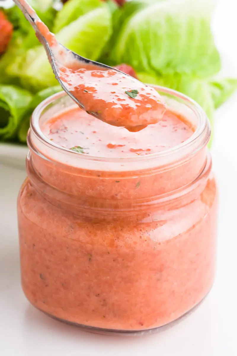 A spoon full of strawberry vinaigrette hovers over a mason jar full of it. There is a green salad behind it.