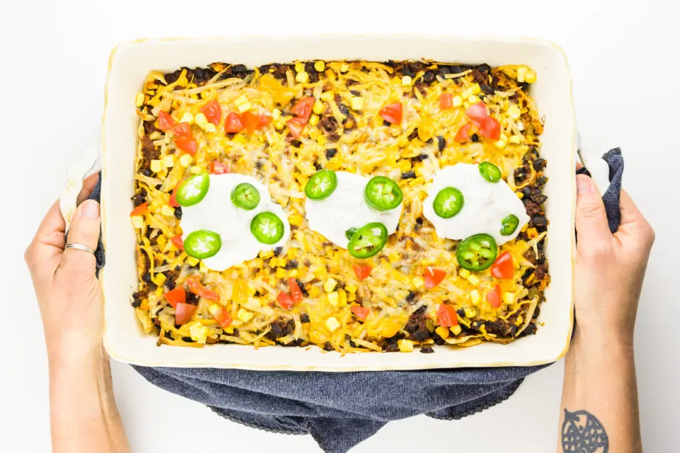 Two hands hold a casserole dish full of freshly baked vegan taco casserole with vegan sour cream on top.