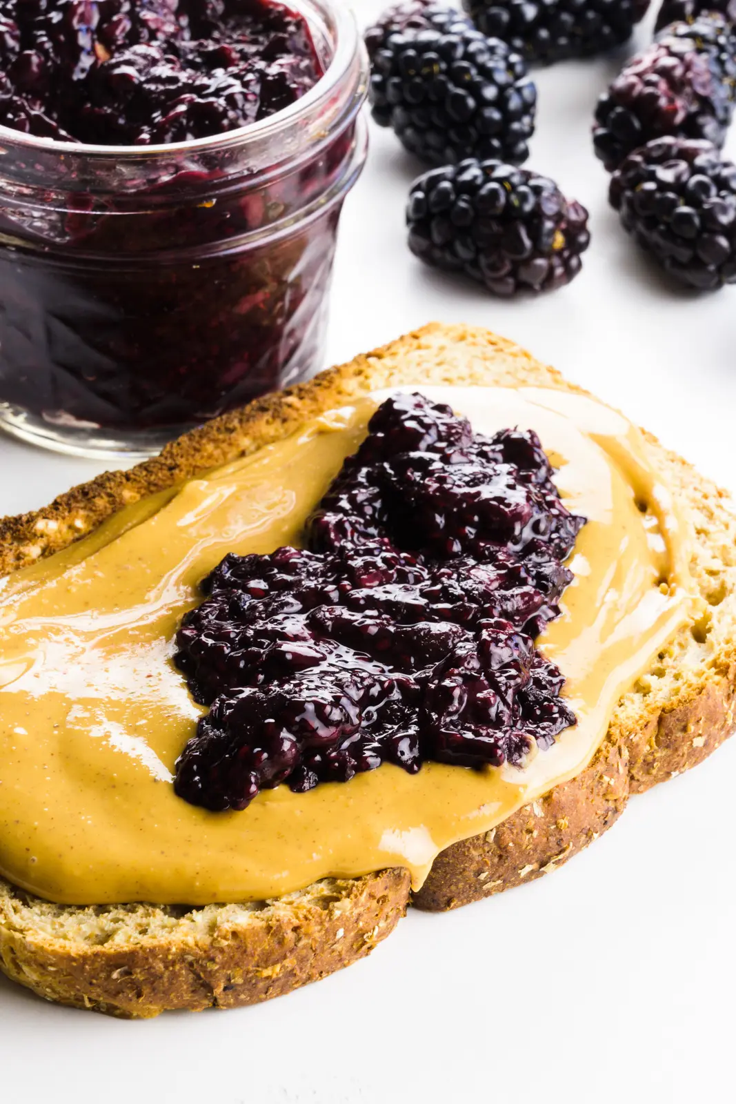 A slice of toast has peanut butter and chia jam on top. There's a jar with more jam behind it and fresh blackberries too.