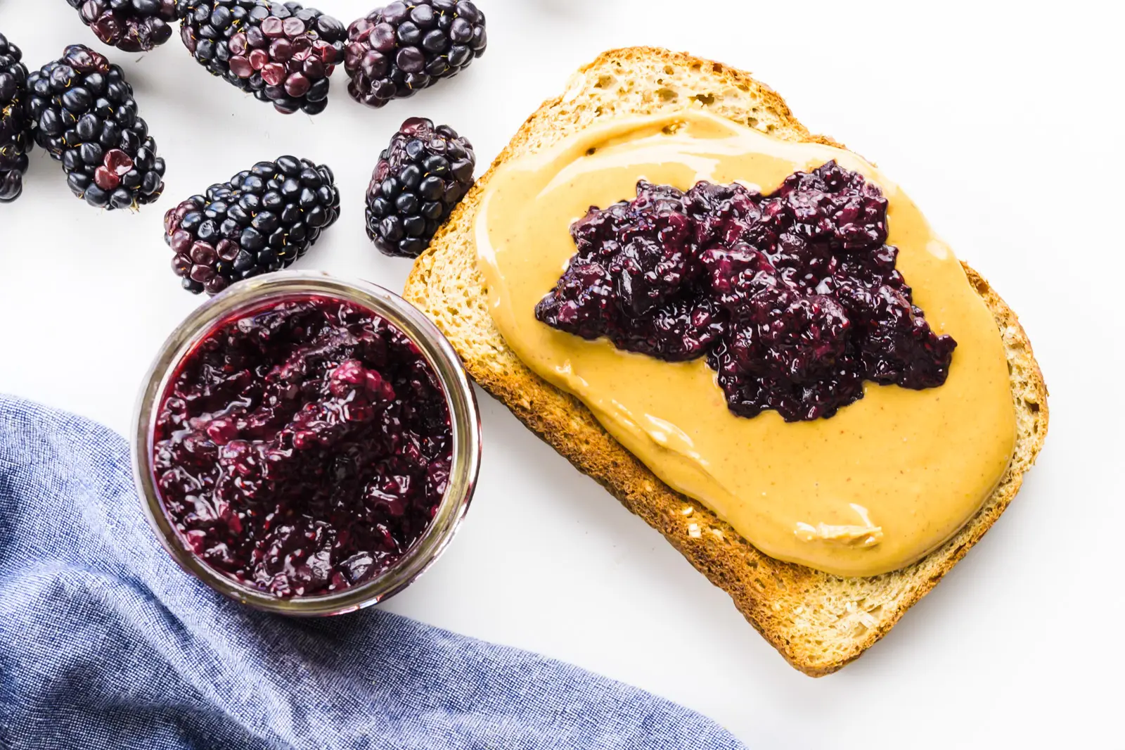 Looking down on a slice of toast with peanut butter and chia jam on top. There's a jar of jam and fresh blackberries beside it.