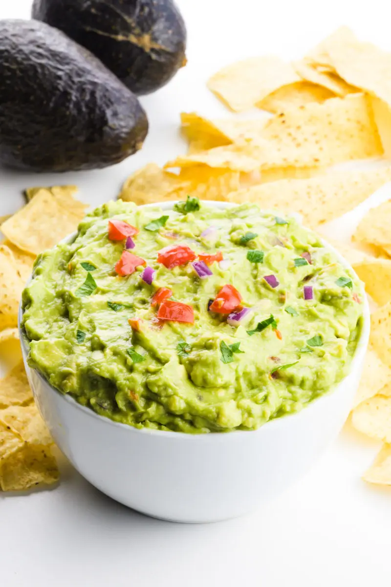 A bowl of vegan guacamole has tortilla chips around it and two avocados in the background.