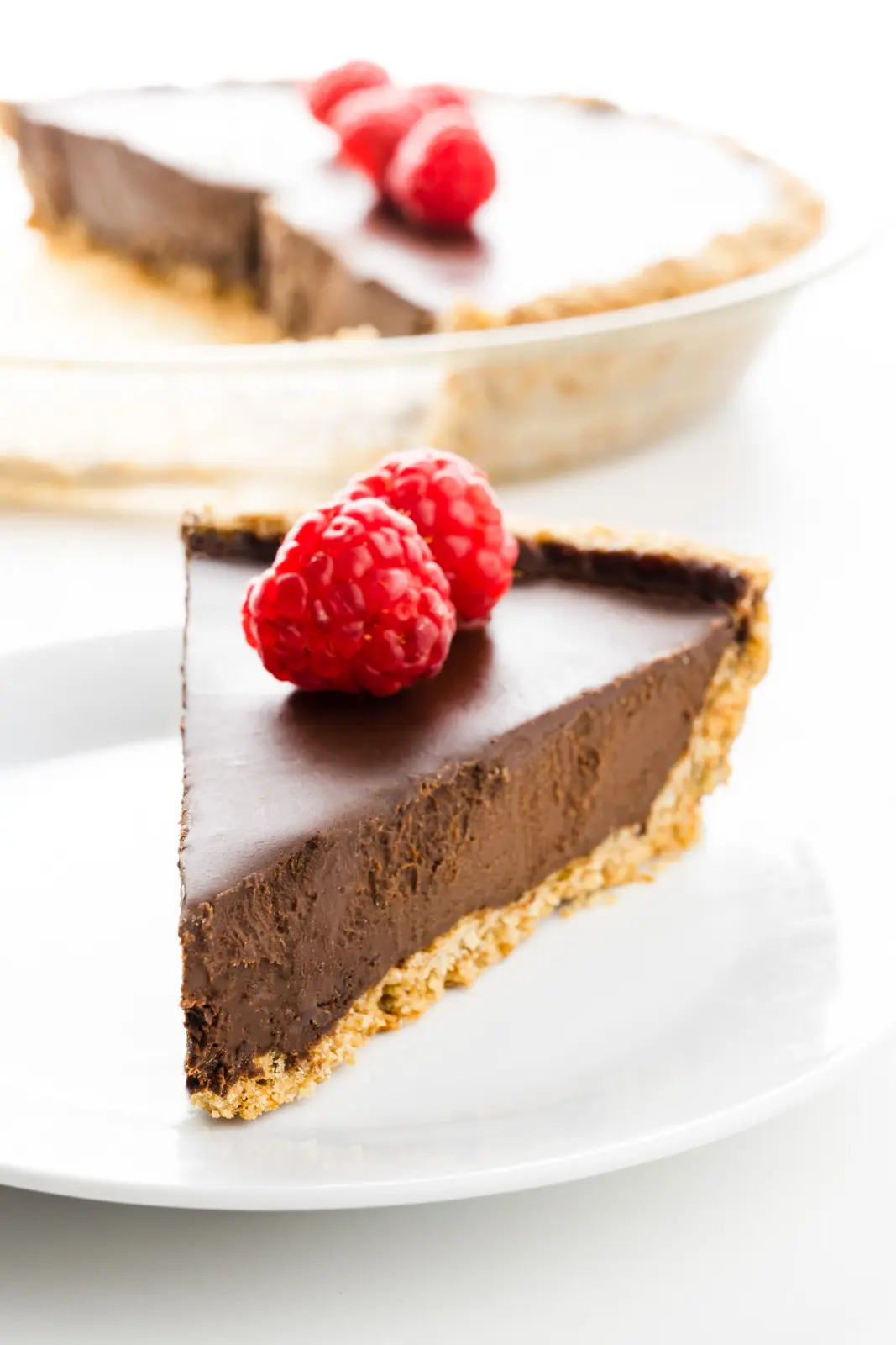 A slice of dark chocolate pie has raspberries on top. The rest of the pie is behind it.