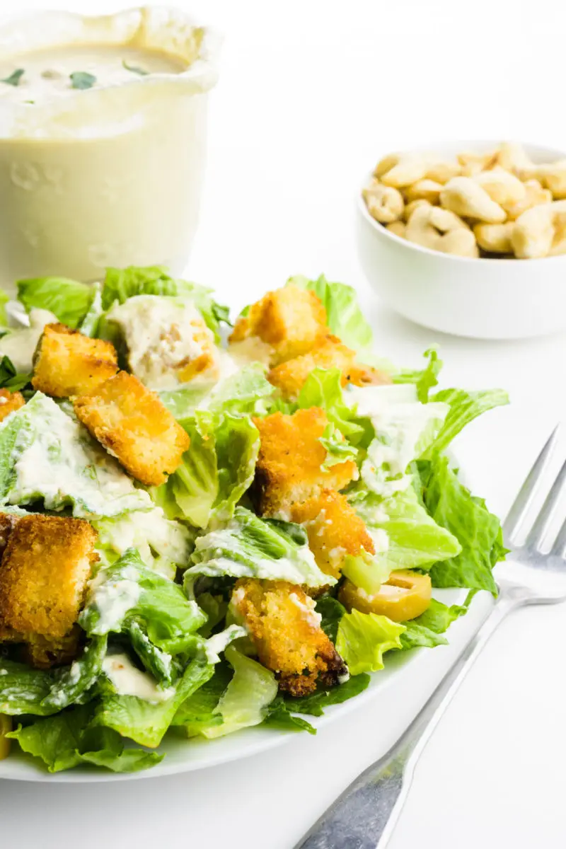 A vegan caesar salad is topped with croutons, olives, and dressing. There's a fork, more dressing, and cashews next to it.