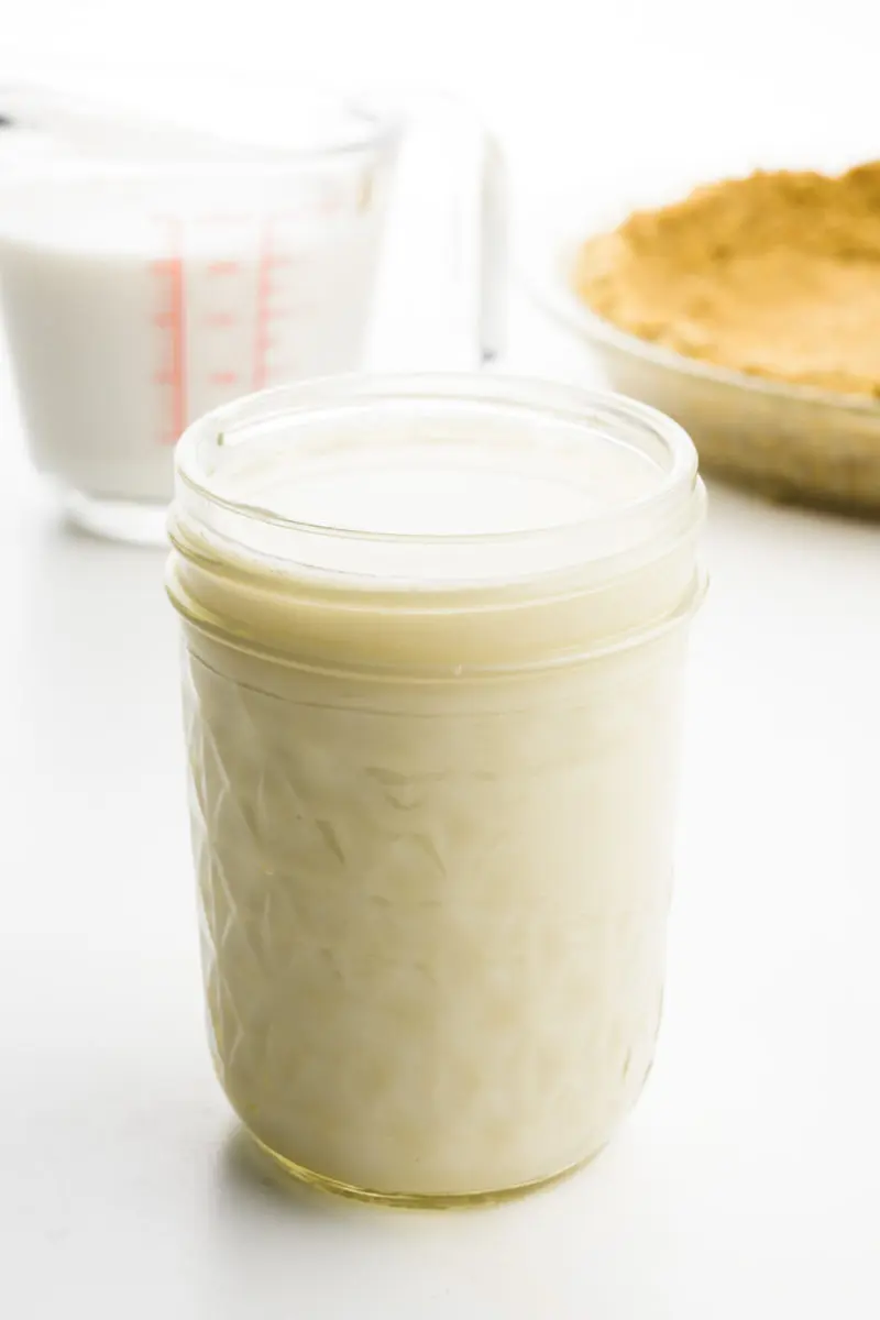 A small mason jar holds vegan evaporated milk. Behind it is a measuring glass with plant-based milk and a pie crust.