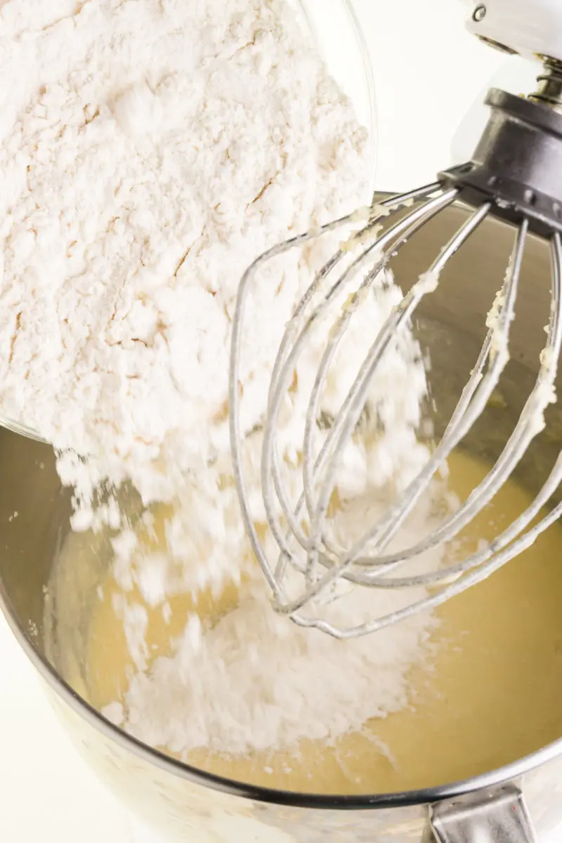 flour is being poured into a mixing bowl of a stand mixer.  There is some batter at the bottom of the bowl.