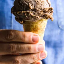 A hand holds a cone full of a large scoop of vegan chocolate ice cream. The person holding it has on a blue top.