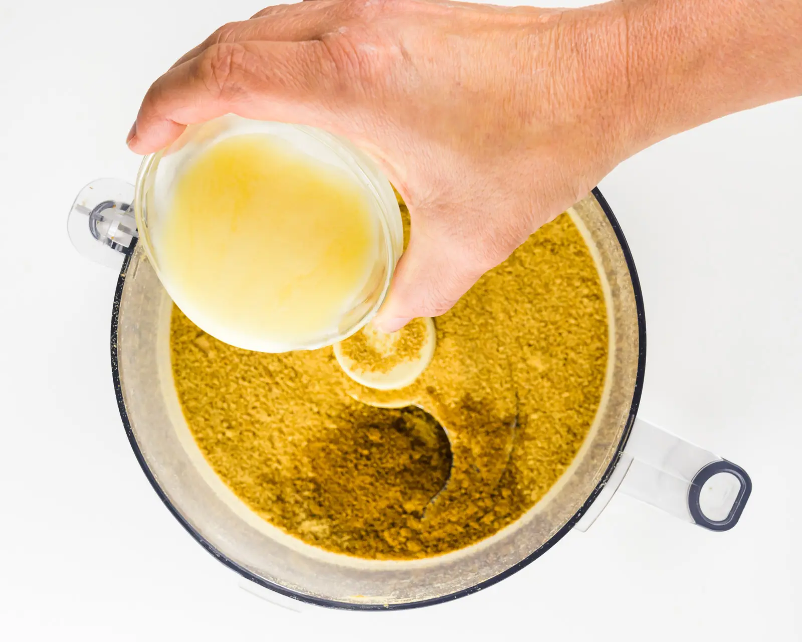 A hand holds a bowl of melted butter and is pouring it into a food processor full of crumbs.
