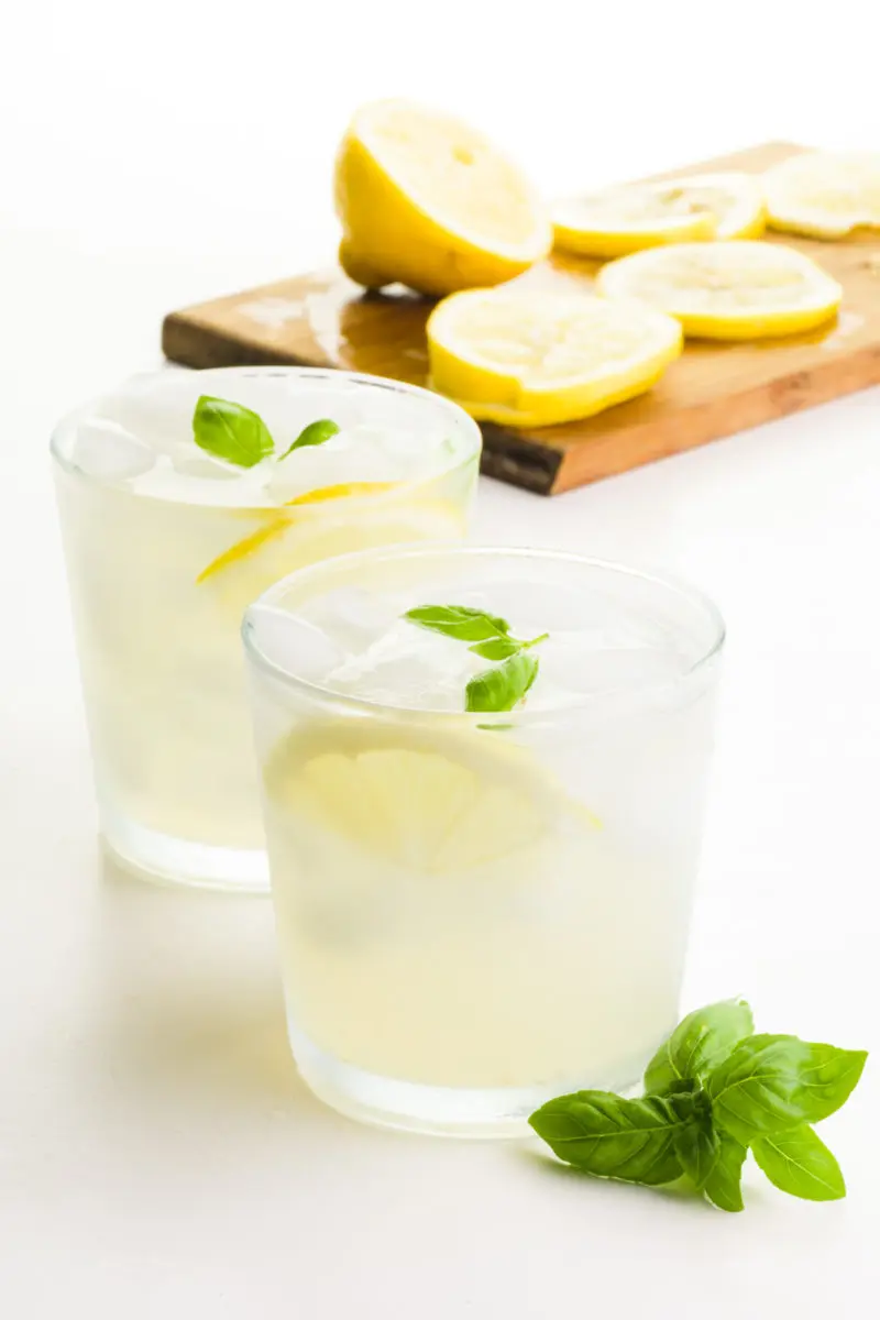 Two glasses hold lemonade. There's sprigs of basil in front of the glass and a cutting board with freshly cut lemons behind it.