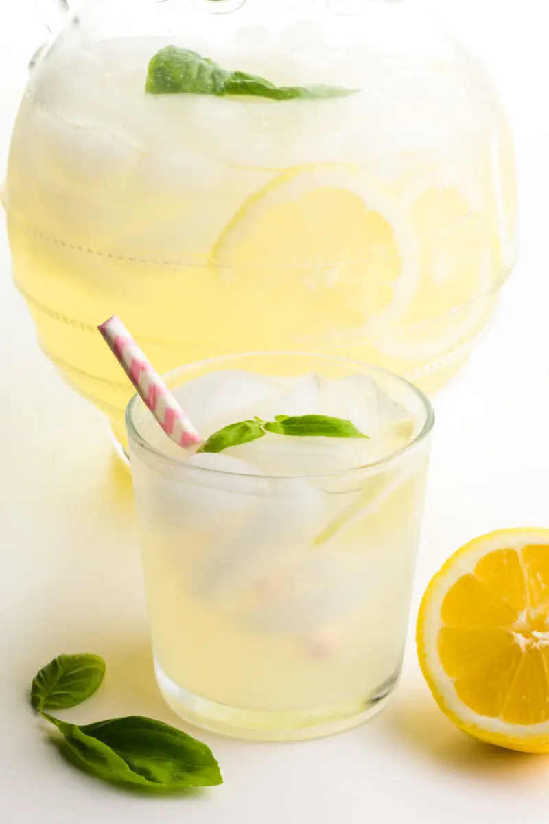 A glass sits in front of a pitcher. Both hold lemonade with fresh lemons and basil leaves.